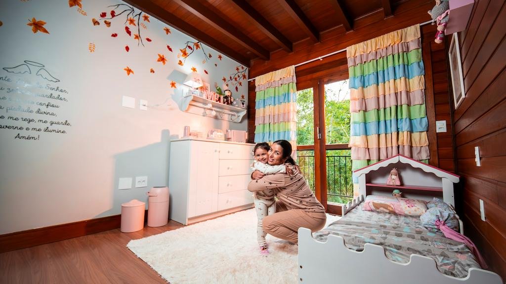 Quarto pequeno com frald&aacute;rio montado sobre c&ocirc;moda branca e m&atilde;e abra&ccedil;ando a filha em um ambiente acolhedor