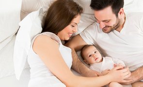 Família com recém-nascido na cama, ilustrando como cuidar de bebê com carinho e segurança no dia a dia