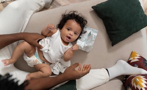 Bebê deitado no sofá recebe troca de fralda, com o pai cuidando e a fralda limpa posicionada ao lado.